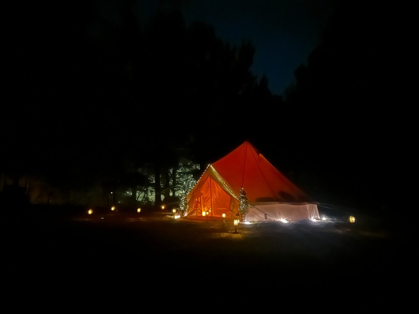 Bell tent at night in a forest clearing with warm lanterns glowing.